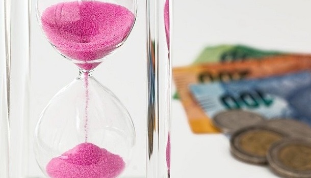 Das Bild zeigt eine laufende Sanduhr mit Geldscheinen und Geldstücken im Hintergrund